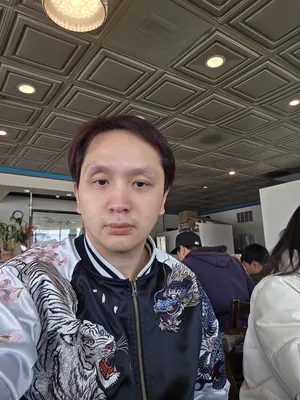 Tommy Nguyen, wearing a black and white embroidered jacket with tiger and dragon designs, takes a self-portrait while sitting at a table in Com Tam Dat Thanh. The photo shows the restaurant's interior, with a casual dining atmosphere.