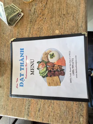 A close-up of the menu cover at Com Tam Dat Thanh, showcasing a colorful dish with grilled meats and rice, placed on a marble table with some money on the side.