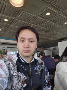 Tommy Nguyen, wearing a black and white embroidered jacket with tiger and dragon designs, takes a self-portrait while sitting at a table in Com Tam Dat Thanh. The photo shows the restaurant's interior, with a casual dining atmosphere.