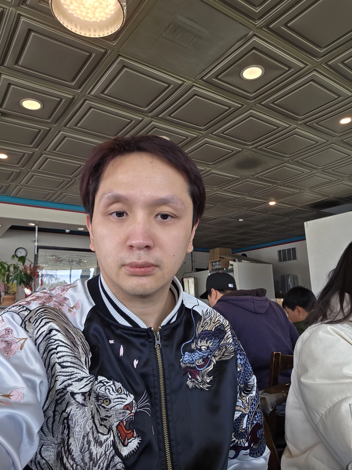 Tommy Nguyen, wearing a black and white embroidered jacket with tiger and dragon designs, takes a self-portrait while sitting at a table in Com Tam Dat Thanh. The photo shows the restaurant's interior, with a casual dining atmosphere.