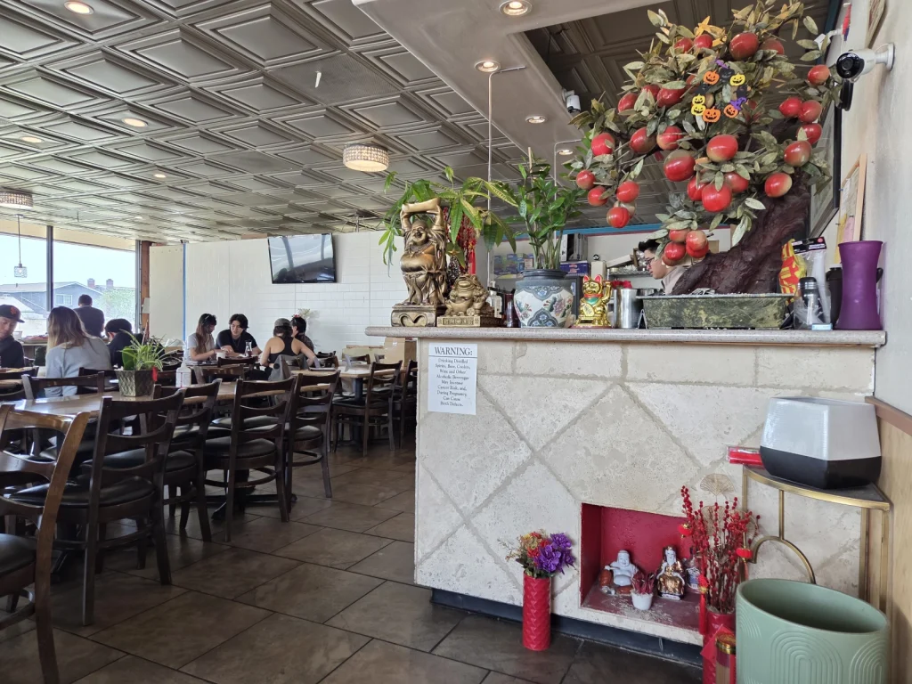 The interior of Com Tam Dat Thanh, featuring a spacious dining area with wooden tables and chairs, decorative plants, and a unique display near the counter.